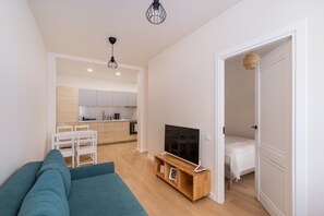 Ein modernes Wohnzimmer mit einer Couch, einem Fernseher auf einem Holzgestell und einer Küche im Hintergrund.