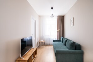 Ein modernes Wohnzimmer mit Fernseher, einer Holzbank, einem blaugrünen Sofa, einem kleinen Beistelltischchen mit einem Ventilator und einem großen Fenster mit durchsichtigen Vorhängen.