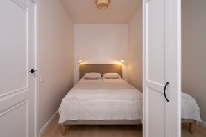 A narrow room with a bed centered, flanked by white doors, a small bedside table, and a wall-mounted light fixture. - Artilerijas 1-bedroom apartment | Courtyard View (Riga)