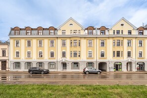 A row of yellow buildings with multiple windows and doors, two cars parked in front, and a grassy area in the foreground. - Artilerijas 1-bedroom apartment | Courtyard View (Riga)