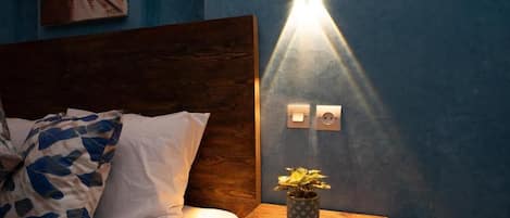 A bedroom with a wooden headboard, a bed with white and blue patterned pillows, a nightstand with a potted plant, and a wall-mounted light fixture.