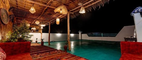 Un área de piscina interior con techo de paja, asientos acolchados rojos y lámparas colgantes.