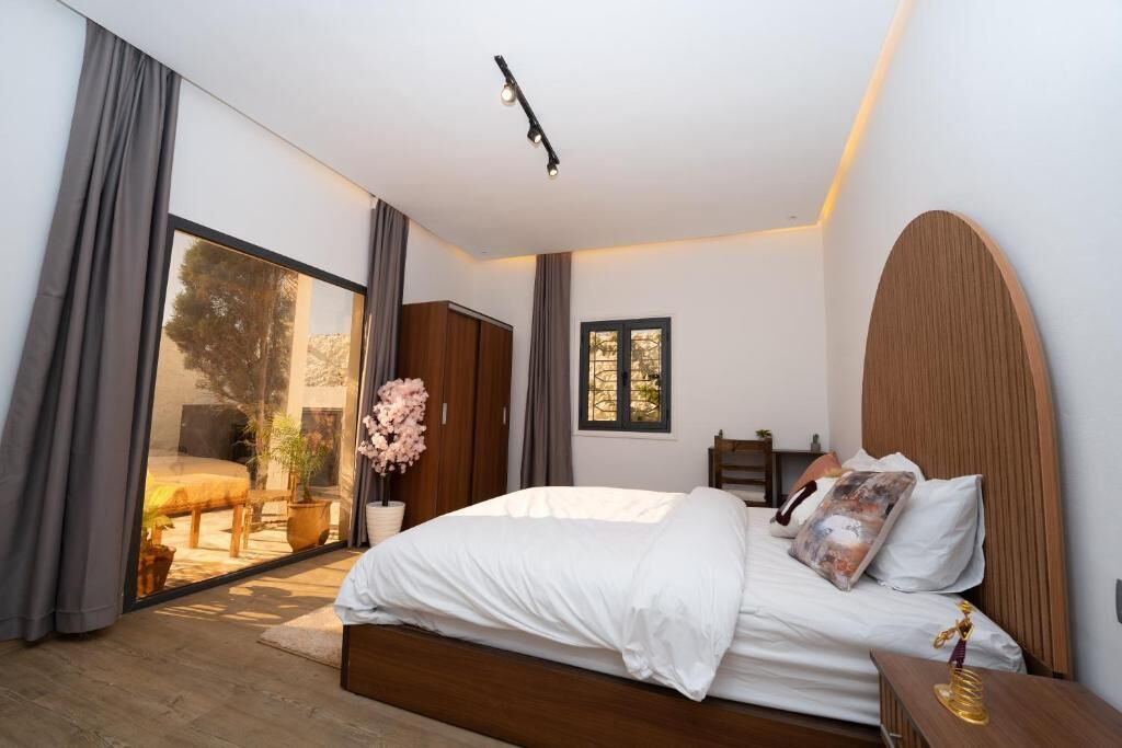 A bedroom with a large bed, a wooden headboard, a bedside table with a lamp, and a view through a sliding door to an outdoor area.