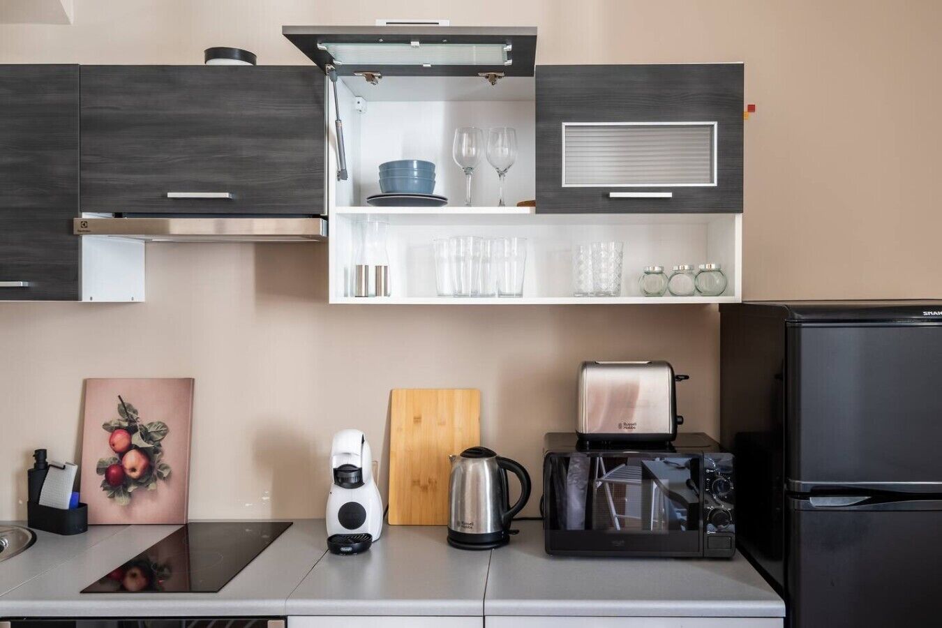 Een moderne keuken met een ingebouwde magnetron, broodrooster en koffiezetapparaat. Op het aanrecht ligt een houten snijplank, een waterkoker en een koffiemachine.