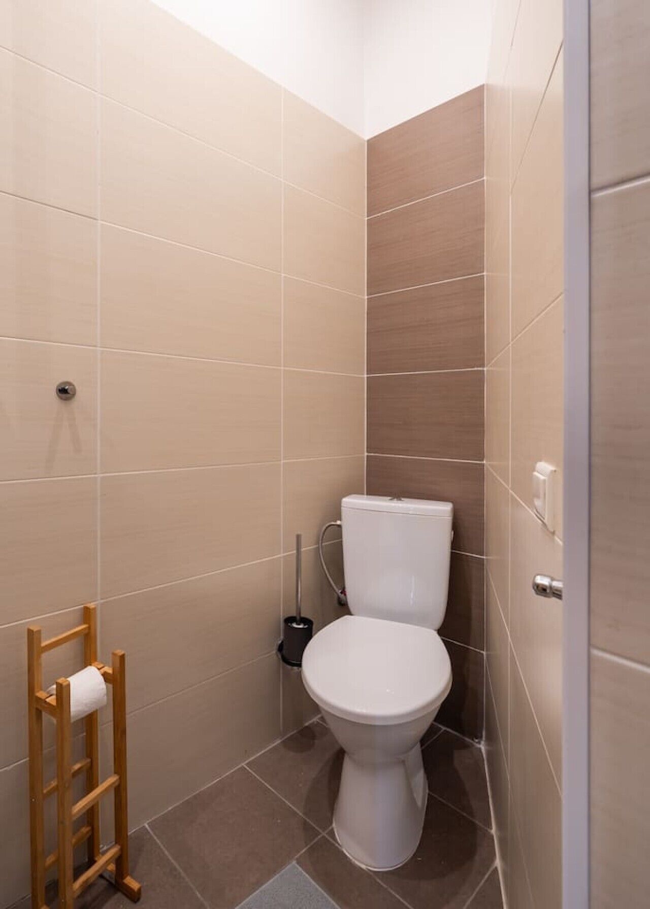 A small, modern bathroom with a toilet, a wooden toilet paper holder, and a plunger on the floor.