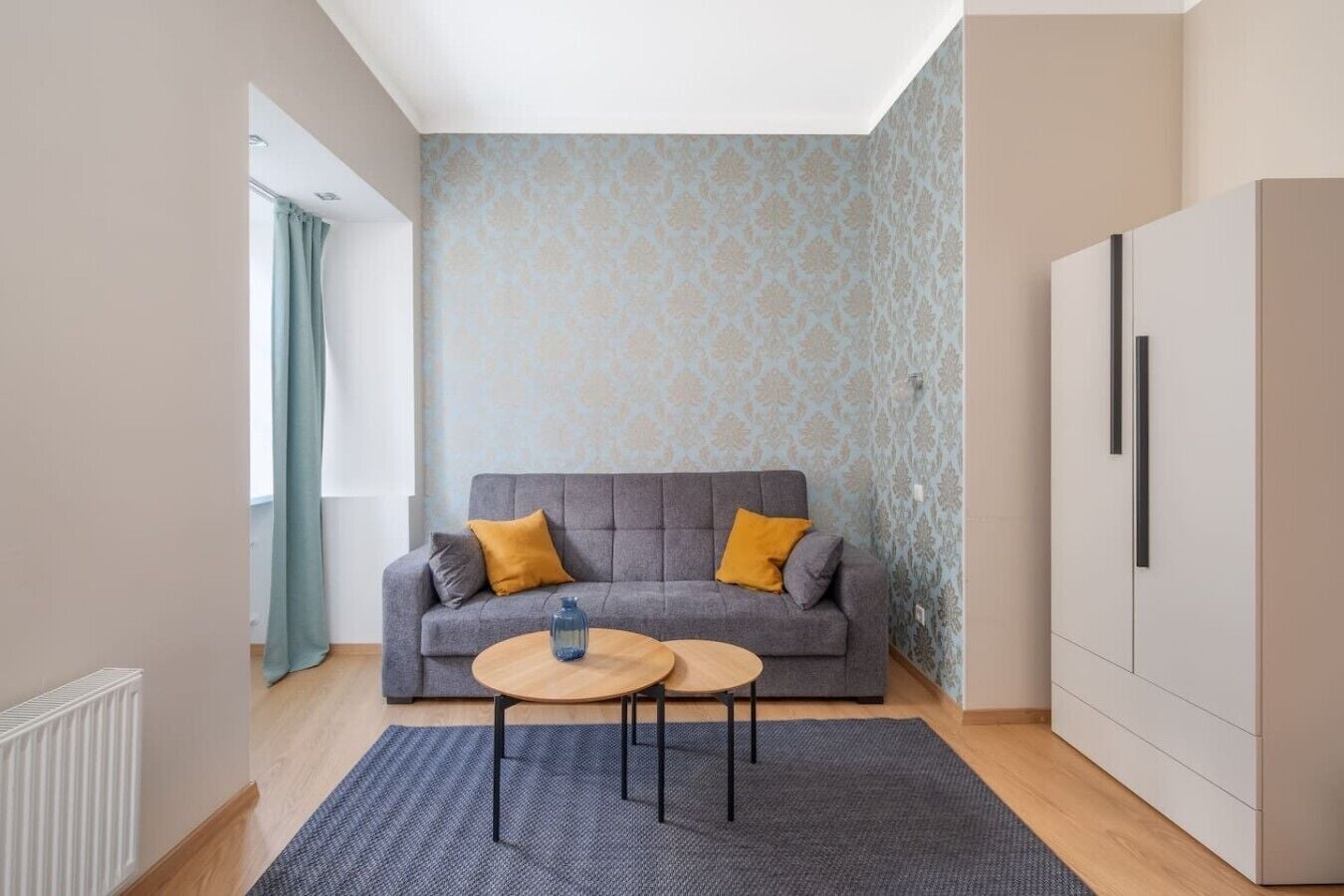 A modern living room with a grey sofa, two round wooden tables, a patterned wallpaper, and a white cabinet.