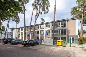 Un edifici de diversos pisos amb zona d'aparcament, arbres i una tanca verda.