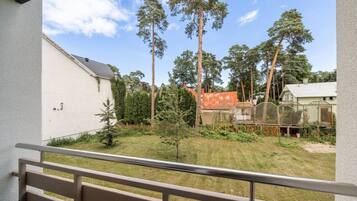 A balcony with a view of a grassy yard, tall trees, and a fenced area with a building in the background.