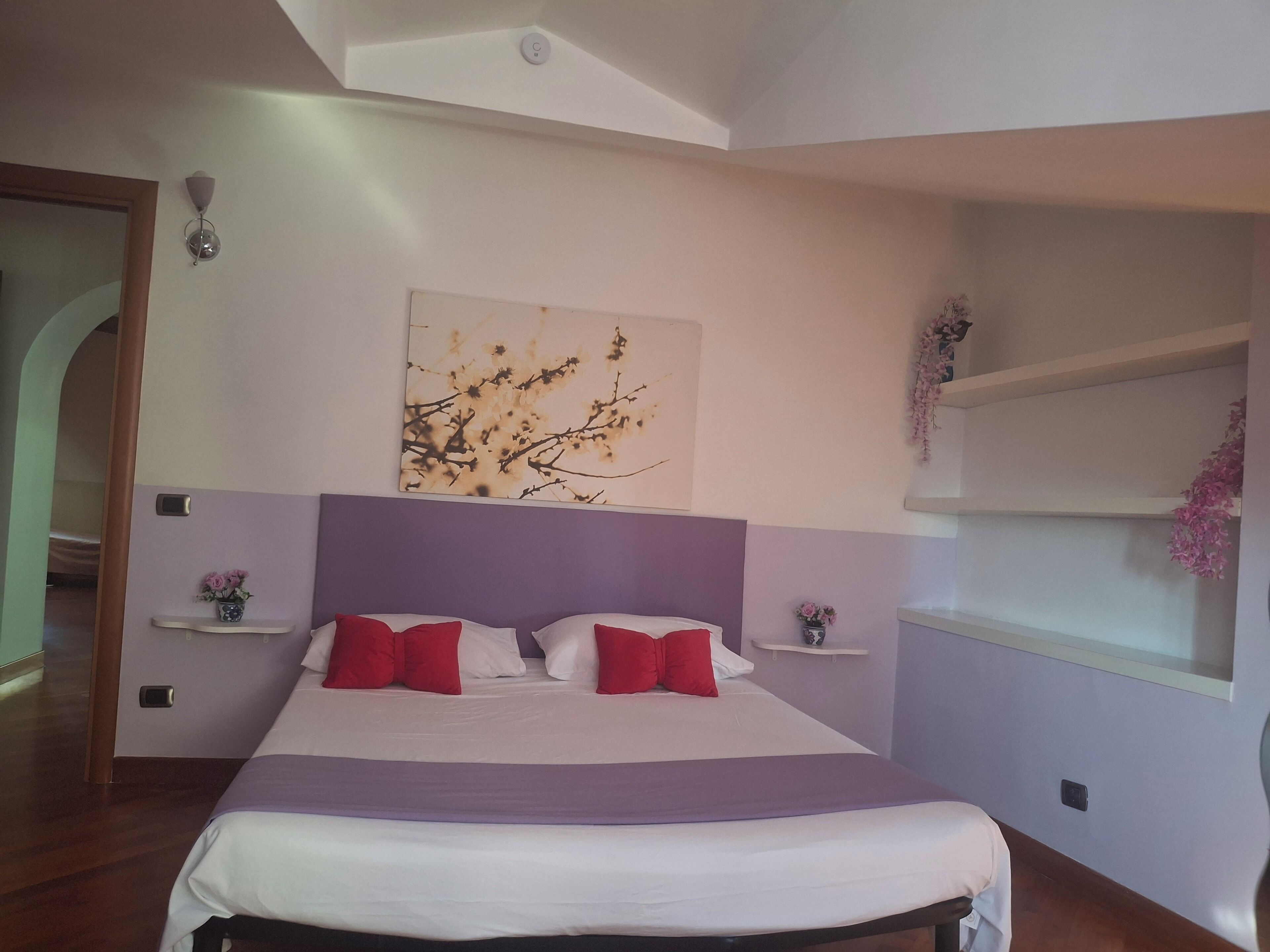 Ein Schlafzimmer mit einem Bett, einem Kopfteil, Nachttischen, einem Wandbild und einem Fenster mit einer Pflanze.