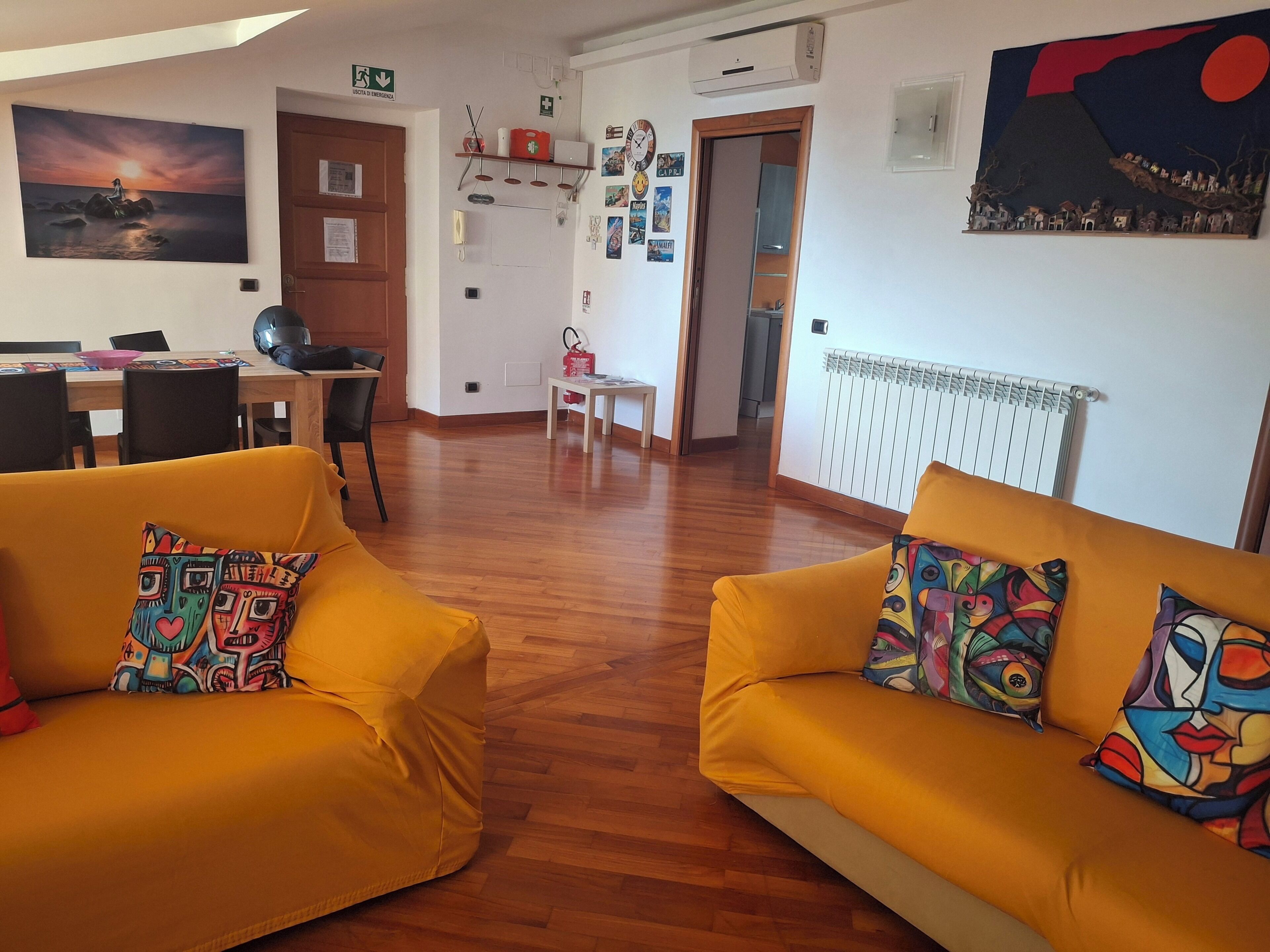 Ein Wohnzimmer mit zwei gelben Sofas, einem Esstisch und einer dahinterliegenden Küchenzeile.