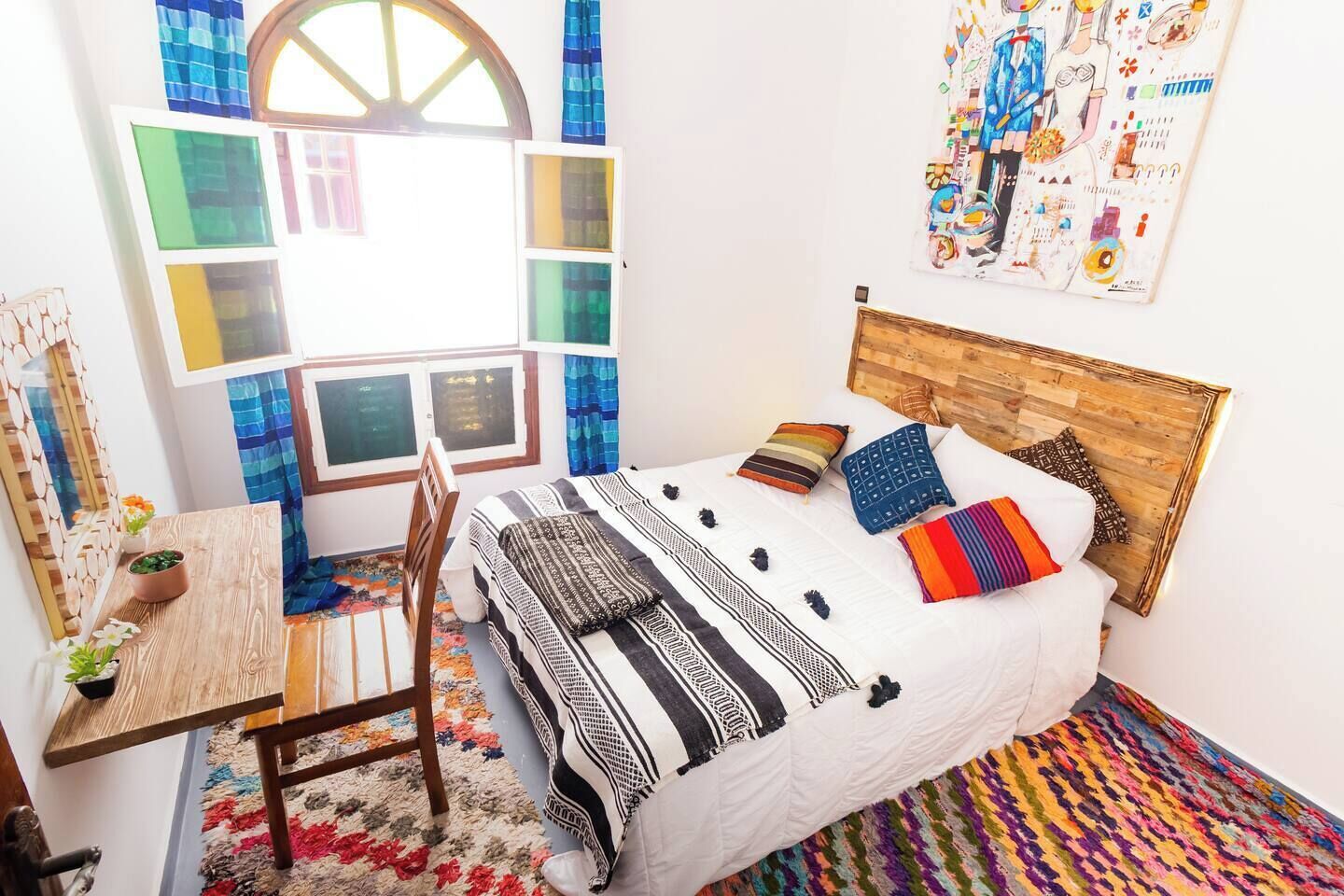 Une chambre avec un lit, un bureau et une chaise. Il y a une fenêtre avec des rideaux, un mur orné d’une affiche et un tapis coloré.