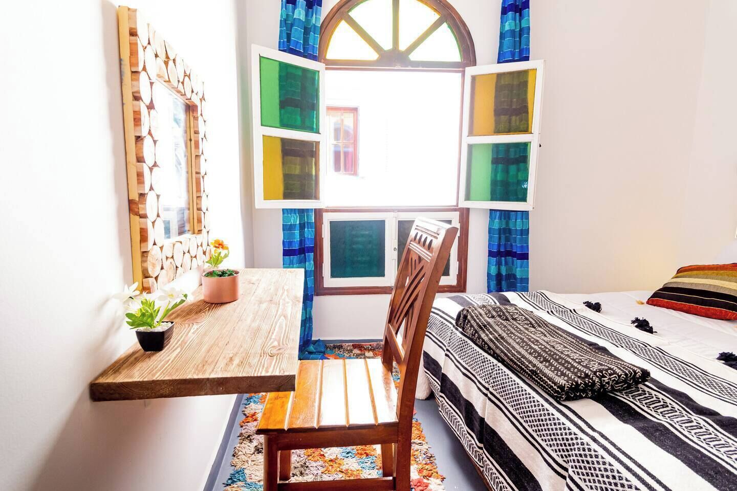 A room with a wooden desk, a chair, a bed with patterned bedding, and a window with colorful curtains.