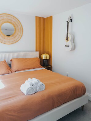 A neatly made bed with orange bedding, a round mirror, a guitar on the wall, and a lamp on a bedside table. - Beautiful modern 2Bdr sunny home with French decor (Hamilton)