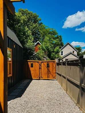 A gravel pathway leading to a wooden gate, with a house and trees in the background. - Beautiful modern 2Bdr sunny home with French decor (Hamilton)