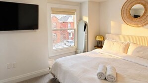 Een slaapkamer met een bed, een flatscreen televisie, een raam, een spiegel en een wandlamp.