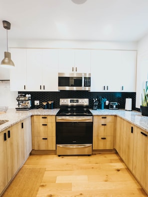 A modern kitchen with white cabinets, a stainless steel microwave, a black stove, and a coffee machine. - Beautiful modern 2Bdr sunny home with French decor (Hamilton)