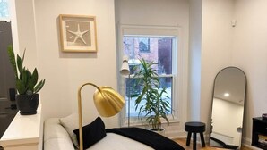 Een moderne woonkamer met een bank, een rond salontafeltje, een vloerlamp, een potplant, een spiegel en een raam met uitzicht.