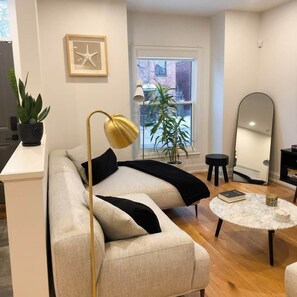 A modern living room with a sofa, a round coffee table, a floor lamp, a potted plant, a mirror, and a window with a view. - Beautiful modern 2Bdr sunny home with French decor (Hamilton)