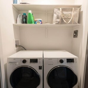 A laundry room with two front-loading washing machines and a shelf above containing cleaning supplies and a laundry bag. - Beautiful modern 2Bdr sunny home with French decor (Hamilton)