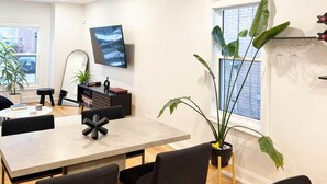 Een moderne woonkamer met een eethoek, een wandgemonteerde flatscreen-tv en een groot raam met uitzicht op de stad.