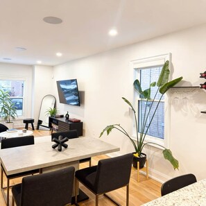 A modern living room with a dining area, a flat-screen TV mounted on the wall, and a large window with a view of the city. - Beautiful modern 2Bdr sunny home with French decor (Hamilton)