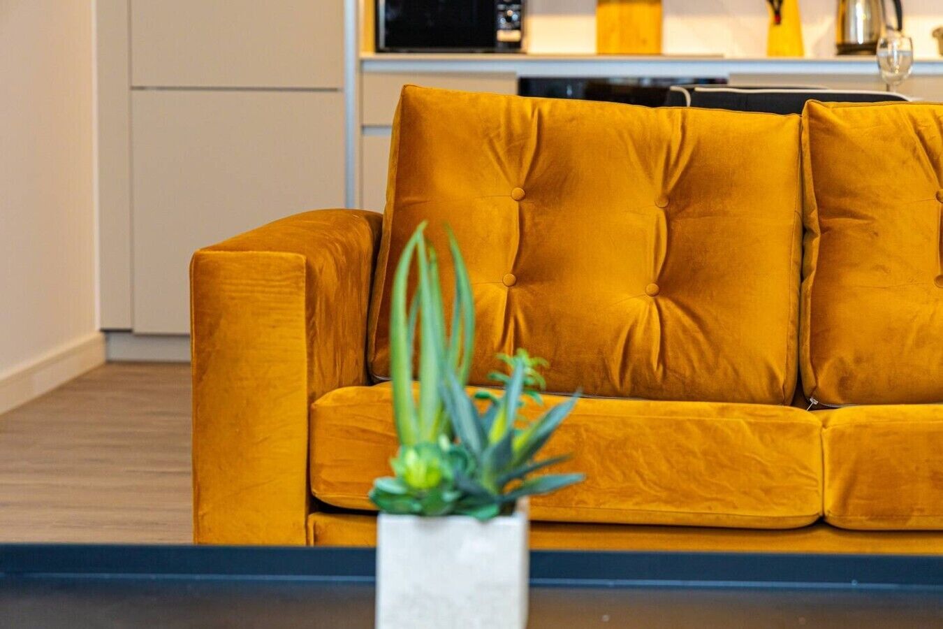 A mustard-colored sofa with a tufted backrest and armrests, positioned in a living area with a white vase containing a plant in the foreground.