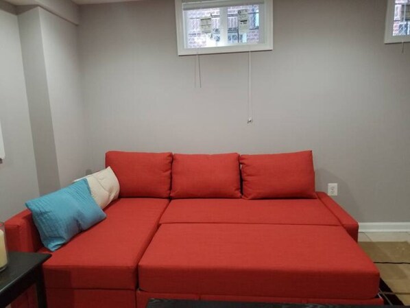 A red sectional sofa with a blue pillow, a small table, and a window with blinds.