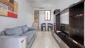A modern living room with a grey sofa, a dining area with chairs, a TV, and a kitchen area with cabinets. - Sliema Central 1BR Apt | Own Entrance, Walk to Sea (Tas-Sliema)