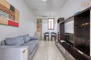 A modern living room with a grey sofa, a dining area with chairs, a TV, and a kitchen area with cabinets. - Sliema Central 1BR Apt | Own Entrance, Walk to Sea (Tas-Sliema)