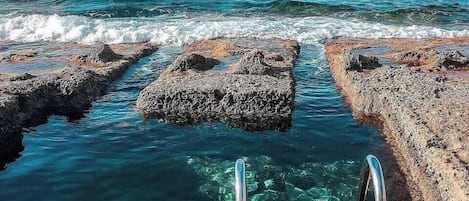 A rocky shoreline with clear water and metal steps leading into the sea.