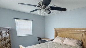 Un dormitorio con cama, cómoda, ventilador de techo, ventana con persianas y piso alfombrado.