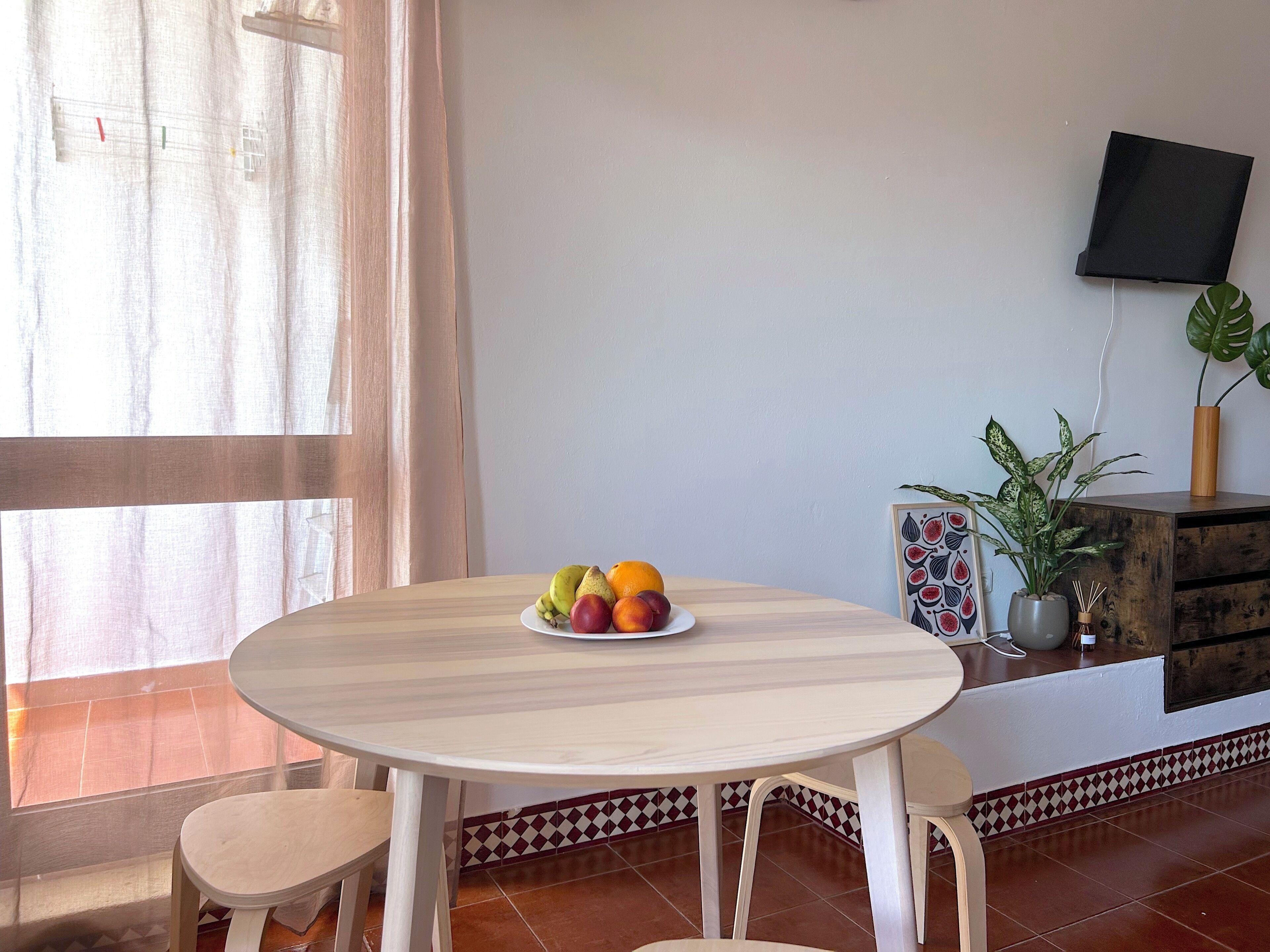 A room with a round table, two chairs, a fruit bowl, a potted plant, a framed picture, and a television on the wall.