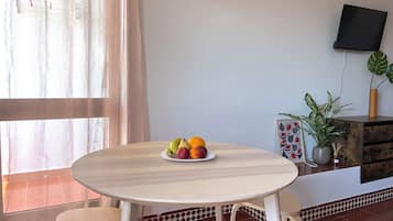 A room with a round table, two chairs, a fruit bowl, a potted plant, a framed picture, and a television on the wall.