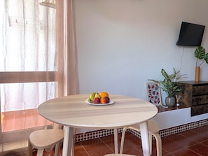 A room with a round table, two chairs, a fruit bowl, a potted plant, a framed picture, and a television on the wall. - Cozy Studio with Pool - The Strip street & Beach (Albufeira)