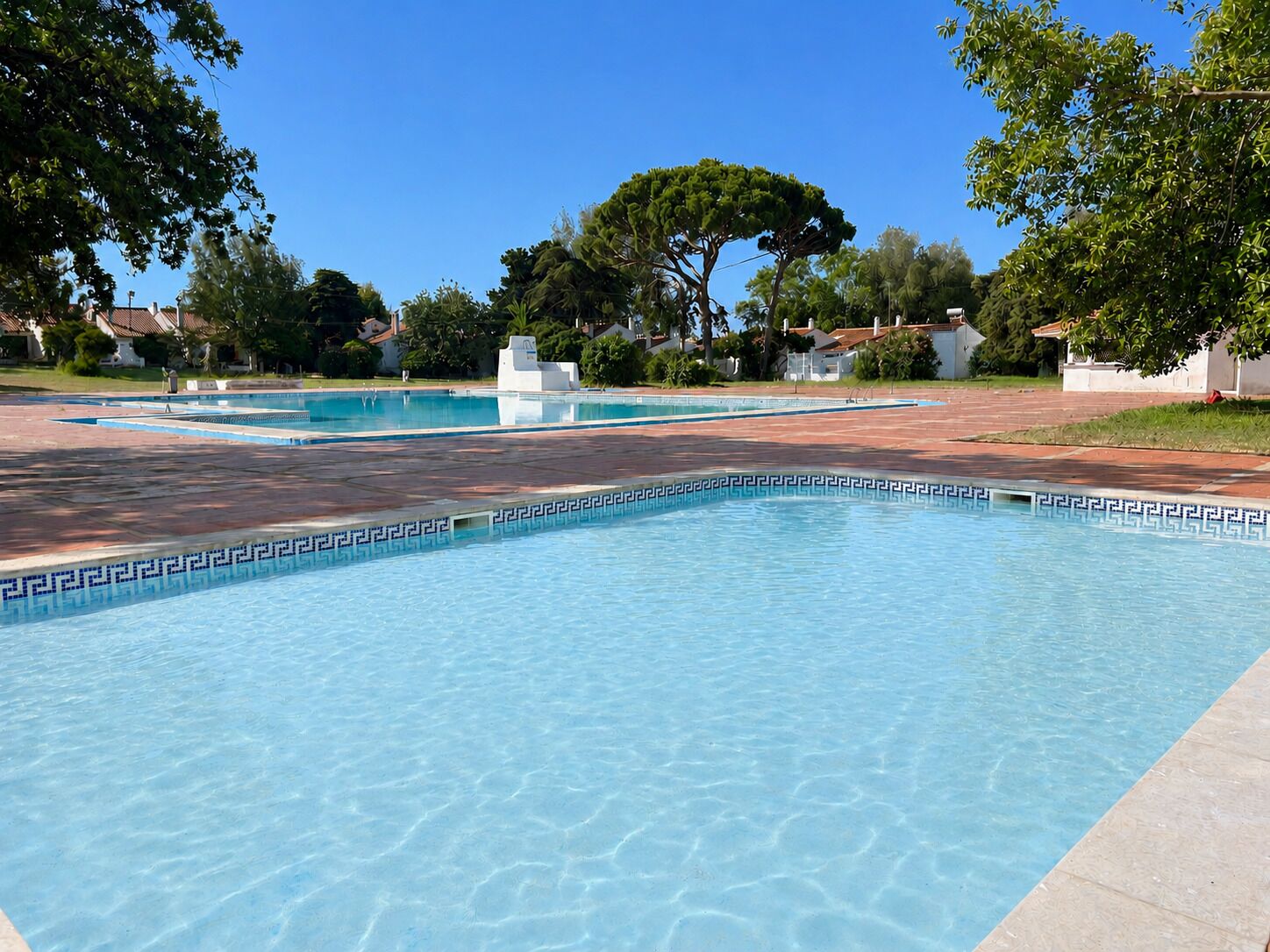 Une piscine aux eaux cristallines, dont le fond est orné de motifs géométriques en mosaïque, entourée d’une zone pavée et d’arbres.