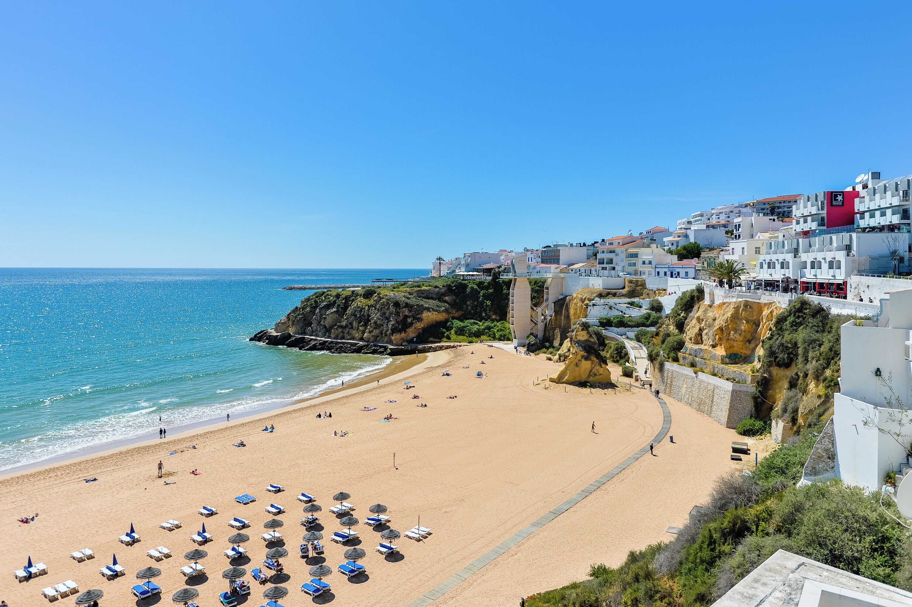 A beach with sun loungers and umbrellas, a clear blue sky, and buildings along the coastline.