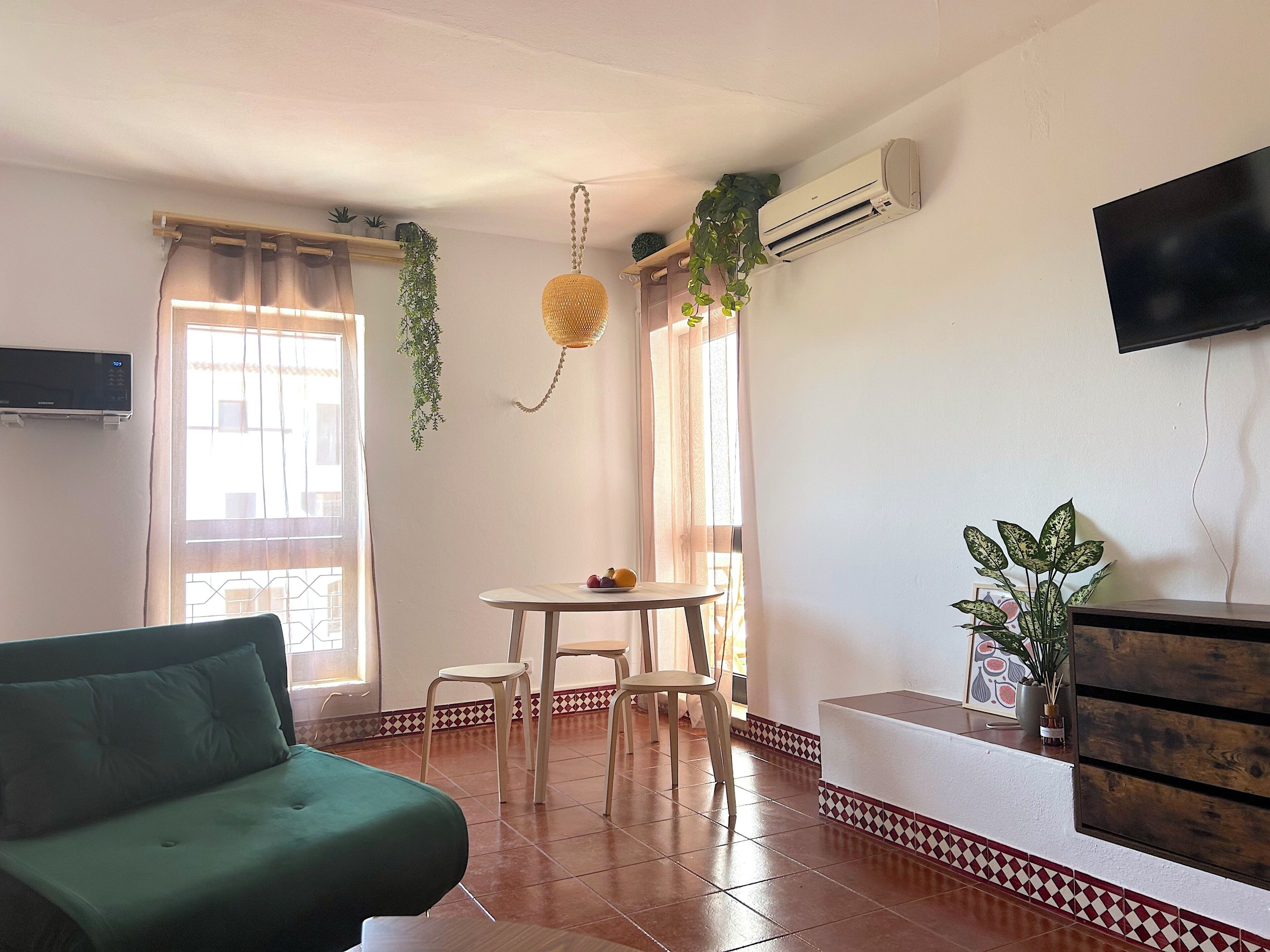 A living room with a green sofa, a dining table with chairs, a TV mounted on the wall, and an air conditioning unit.