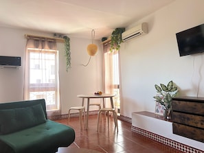 A living room with a green sofa, a dining table with chairs, a TV mounted on the wall, and an air conditioning unit. - Cozy Studio with Pool - The Strip street & Beach (Albufeira)