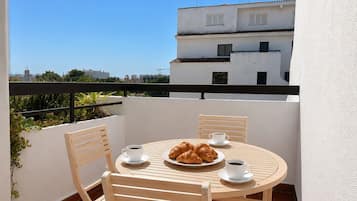 A round wooden table with four chairs, a plate of croissants, and two cups of coffee on a balcony with a white wall and a black railing.