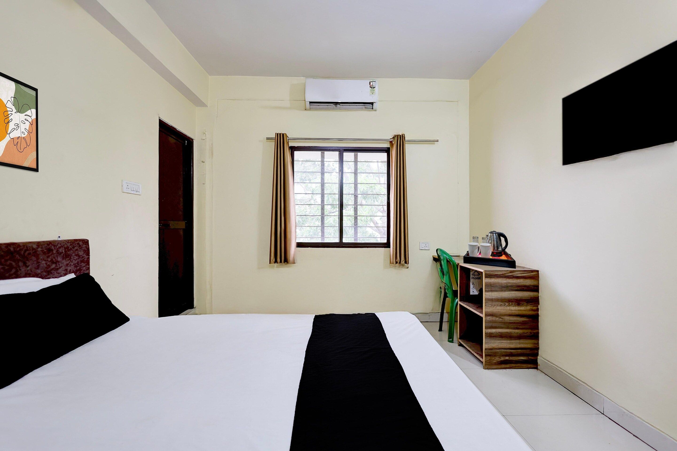 A hotel room with a bed, a window with curtains, a wall-mounted air conditioner, and a small table with a kettle and cups.
