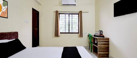 A hotel room with a bed, a window with curtains, a wall-mounted air conditioner, and a small table with a kettle and cups.