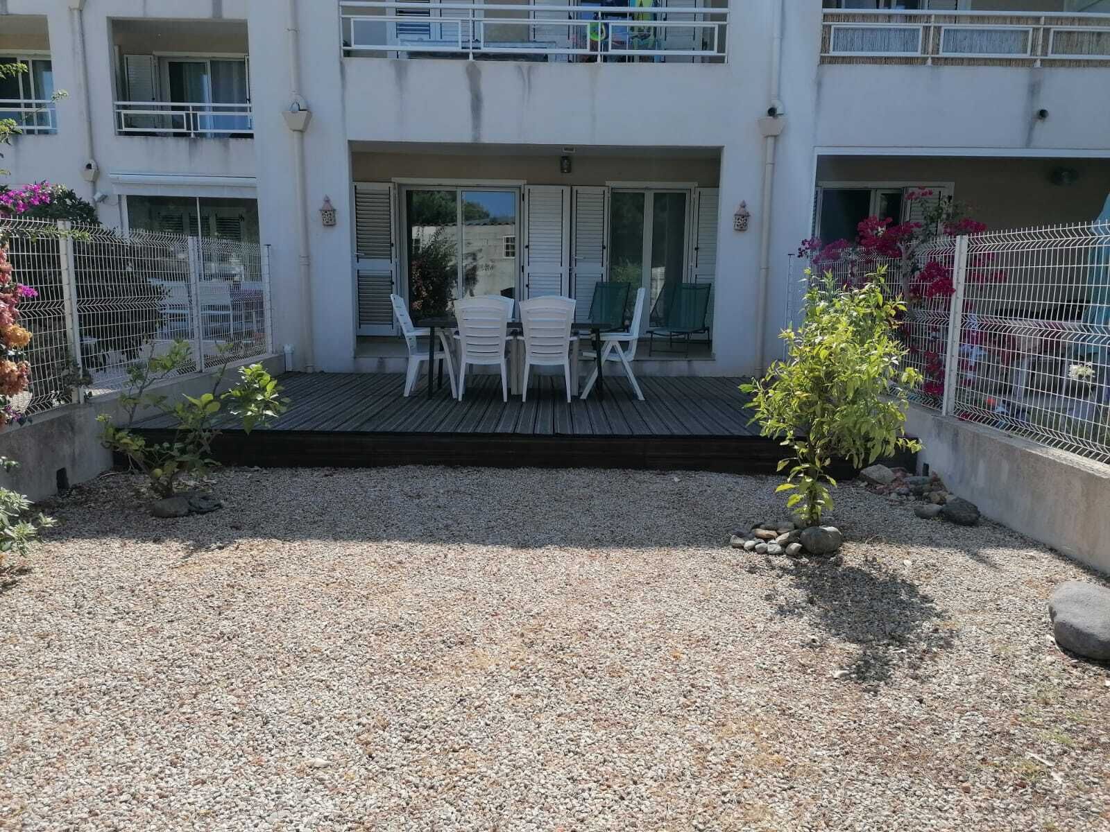 Un bâtiment à deux étages doté d’un espace extérieur couvert avec des sièges, d’une terrasse en gravier et d’un petit jardin agrémenté de plantes et de fleurs.