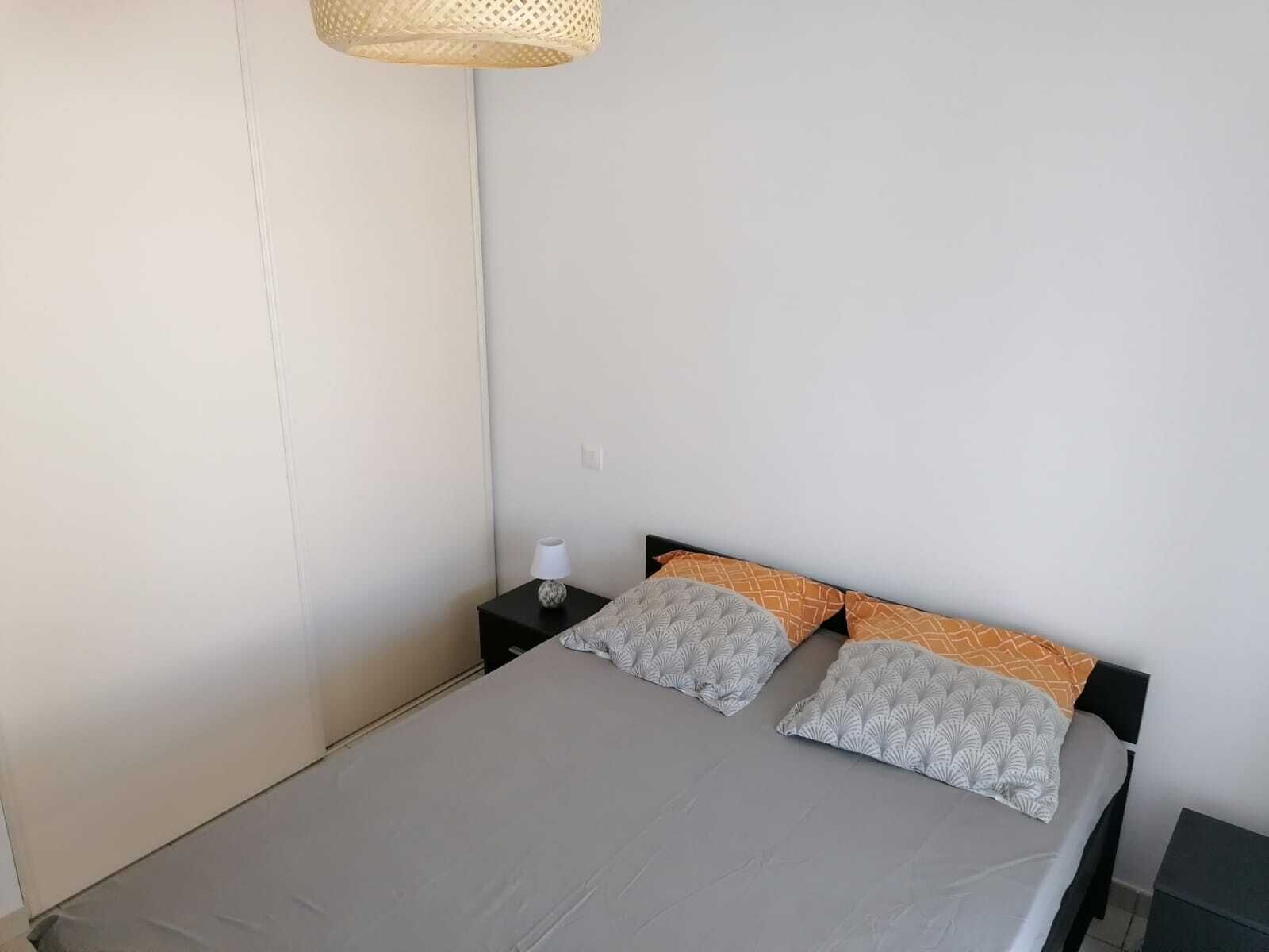 A simple, uncluttered bedroom with a bed, two pillows, a bedside table with a lamp, and a hanging light fixture.
