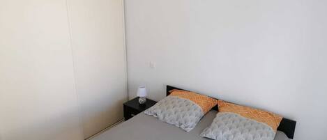 A simple, uncluttered bedroom with a bed, two pillows, a bedside table with a lamp, and a hanging light fixture.
