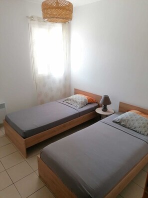 Una habitación con dos camas individuales, una mesita con lámpara, una ventana con cortinas translúcidas y una lámpara de techo de fibras naturales.