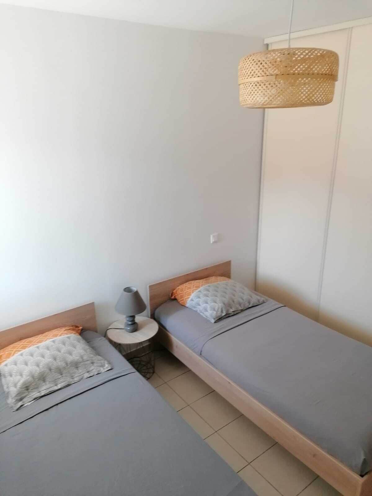 A small, compact room with two single beds, a bedside table with a lamp, and a woven hanging light fixture.