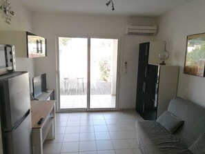 Una sala moderna con sofá, televisor, zona de cocina con refrigerador y puerta corrediza de vidrio que da a un patio exterior.