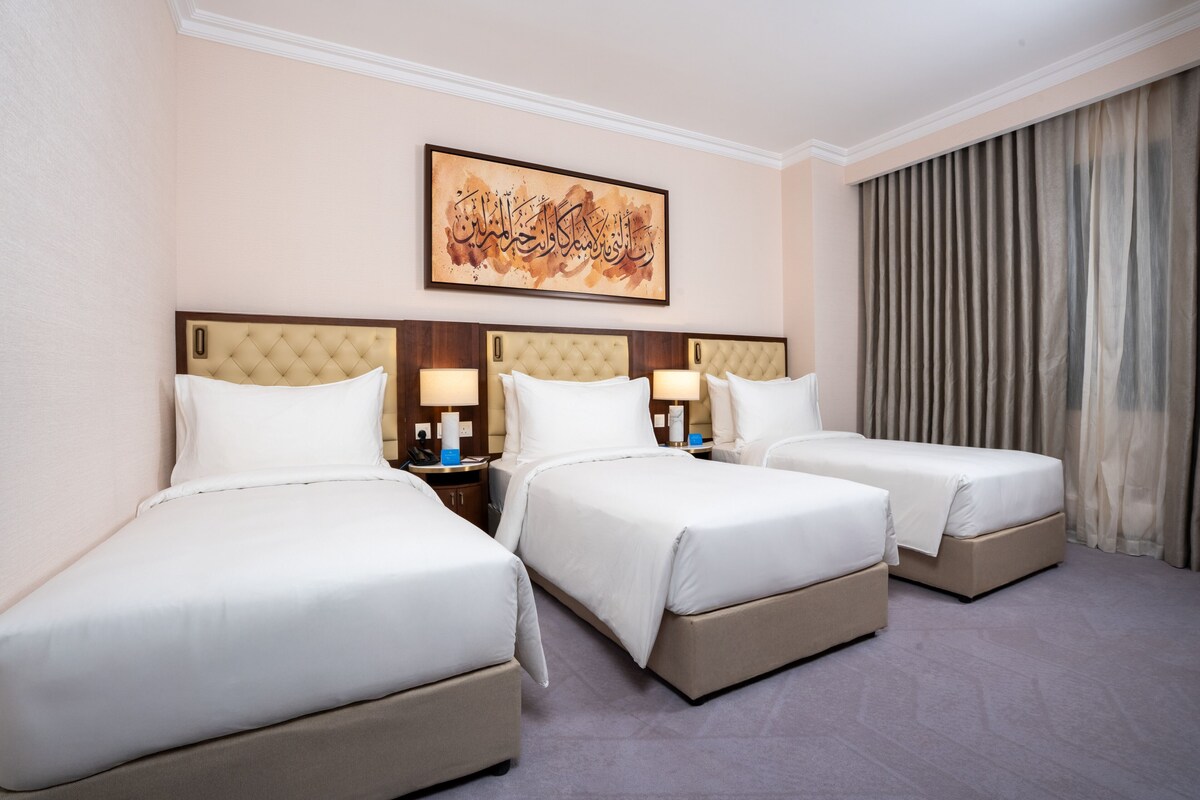 Habitación de hotel con tres camas, una obra de arte grande en la pared y una ventana con cortinas.
