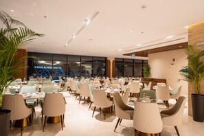 En hotellrestaurang med flera bord och stolar, en modern inredning och utsikt över hotellets fasad.
