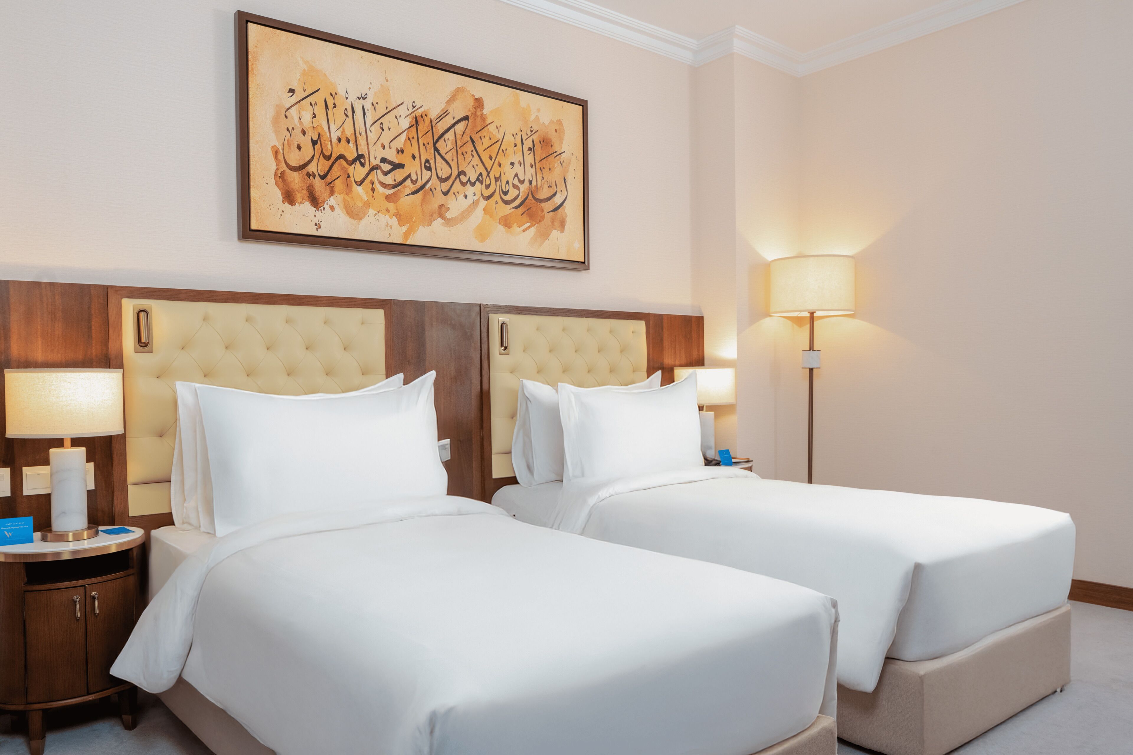 A hotel room with two beds, a nightstand with a lamp, and a wall-mounted artwork.